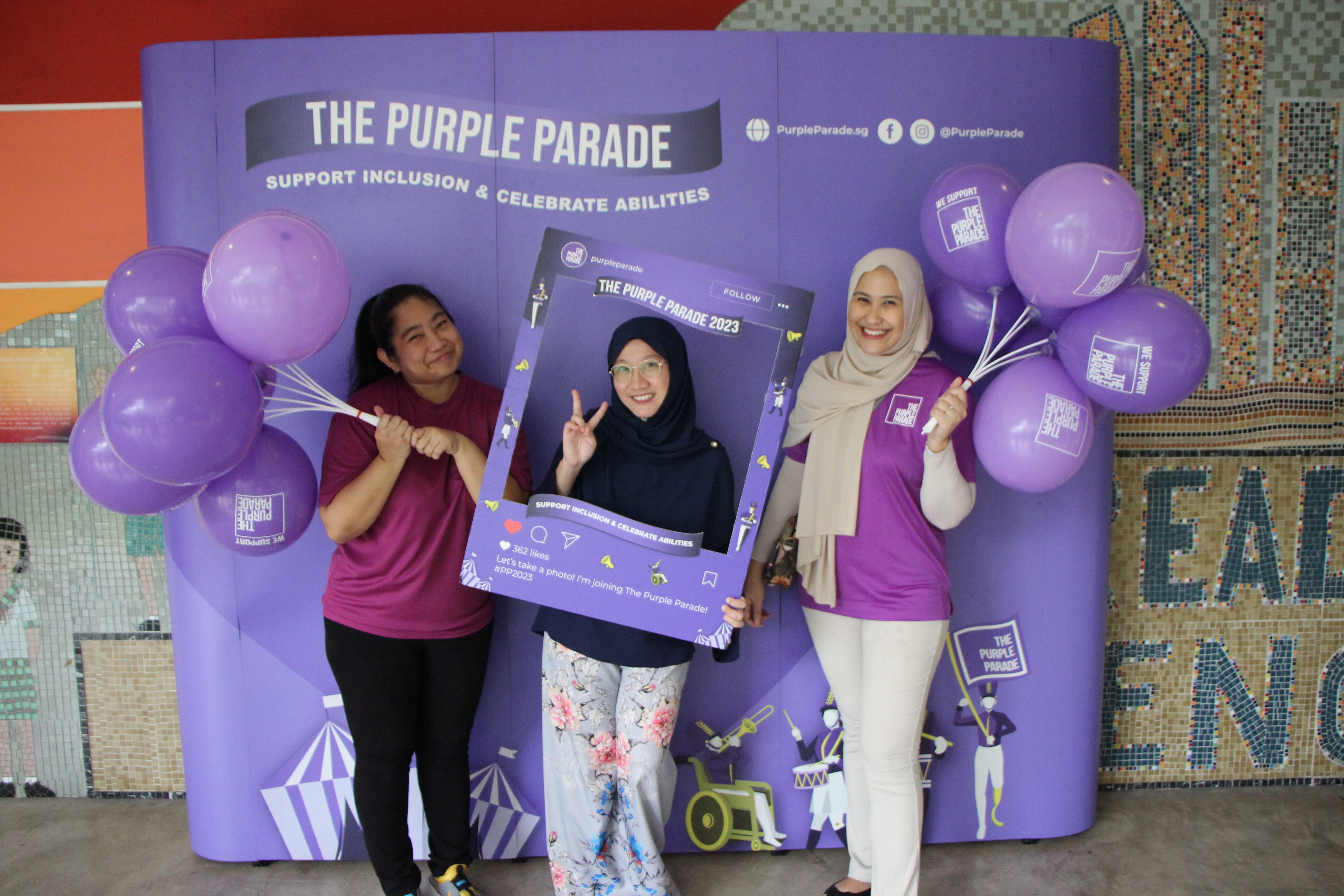 Three women pose with purple balloons and a photo frame in front of The Purple Parade 2023 banner.